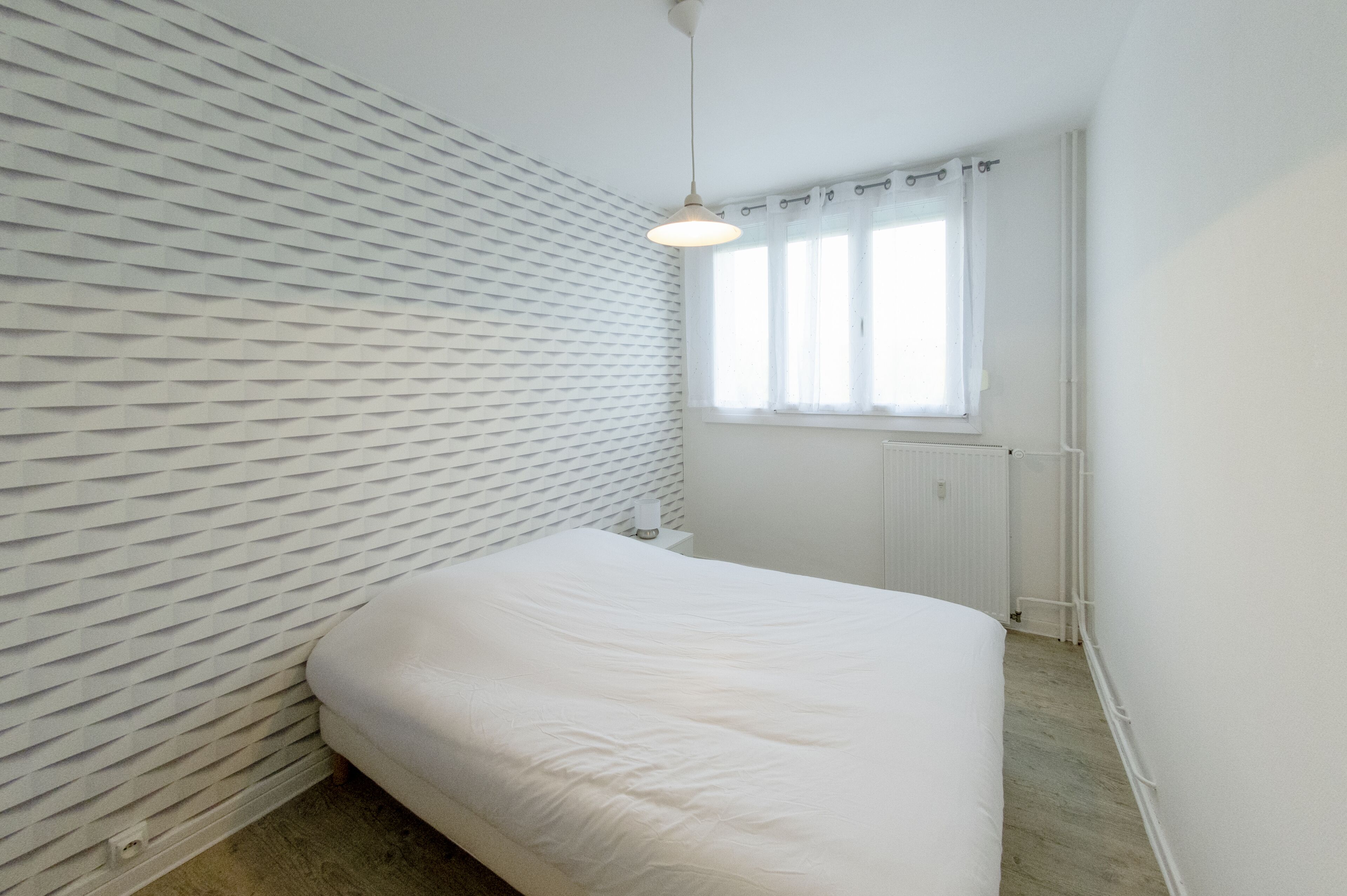 Quarto com cama, janela com cortinas, radiador e parede texturizada.