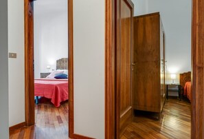 A room with a wooden wardrobe, a bed with a red bedspread, and a nightstand with a lamp.