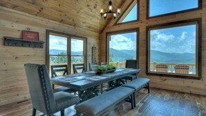 Une cabane en bois dotée d’un coin repas comprenant une table, des chaises et des bancs. De grandes fenêtres offrent une vue sur les montagnes.