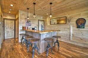 A rustic kitchen with wooden walls, a central island with stools, pendant lighting, and a dartboard.