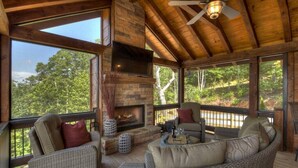Une terrasse en bois avec des meubles en osier, une cheminée et de grandes fenêtres donnant sur une forêt.