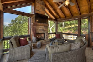 A wooden deck with wicker furniture, a fireplace, and large windows overlooking a forest.