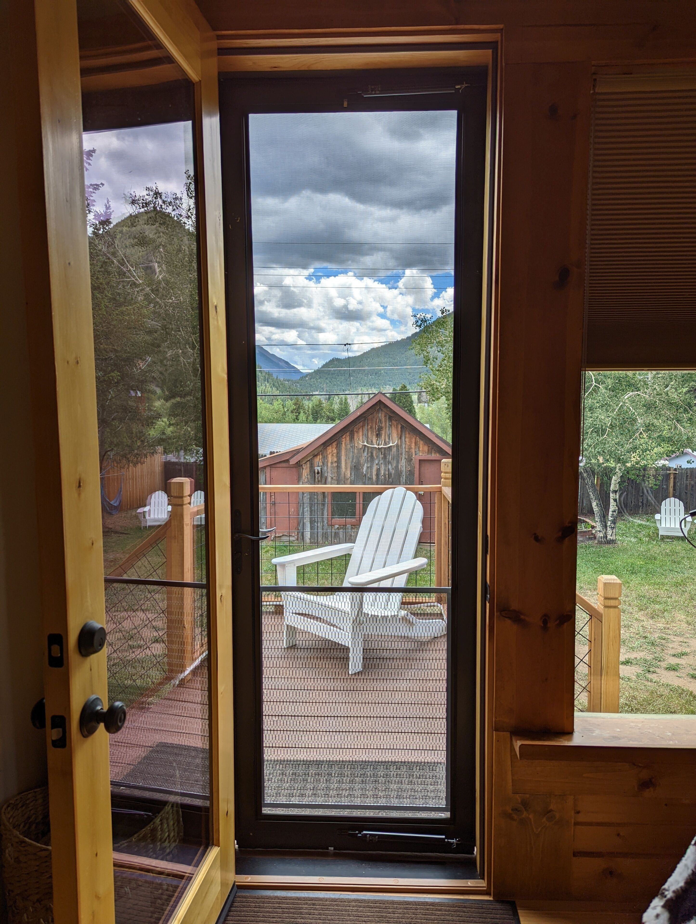 Une porte en bois avec un panneau de verre qui donne sur une terrasse où se trouve une chaise Adirondack blanche, et on aperçoit une cabane en bois au fond.