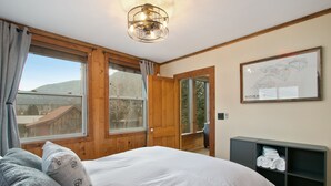 A bedroom with a large bed, wooden walls, a wooden door, a window with a view of a mountain, and a framed map on the wall.