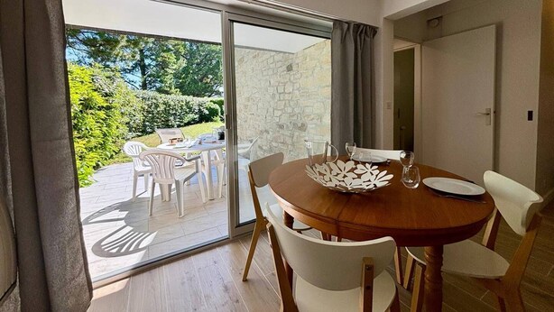 A dining area with a wooden table, white chairs, and a glass door leading to an outdoor patio with a stone wall and greenery. - CARNAC - Apartment 2 Rooms - 2/4 persons (CARNAC)