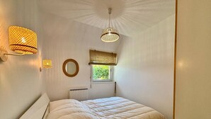 A bedroom with a single bed, two wall-mounted lamps, a round mirror, a window with a view of greenery, and a radiator. - CARNAC - Apartment 2 Rooms - 2/4 persons (CARNAC)