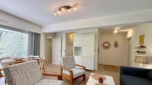 A modern living room with a sofa, armchair, and coffee table. There is a dining area with a table and chairs, and a kitchen area with cabinets. - CARNAC - Apartment 2 Rooms - 2/4 persons (CARNAC)