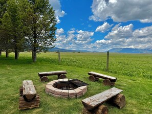 A fire pit with benches in a grassy field with trees and mountains in the background. - Black Bear Meadows - only 22 miles to West Yellowstone! (Island Park)