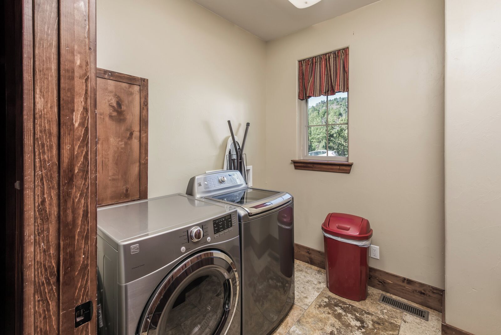 A laundry room with a front-loading washer and dryer, a red trash can, and a window with a view of greenery.