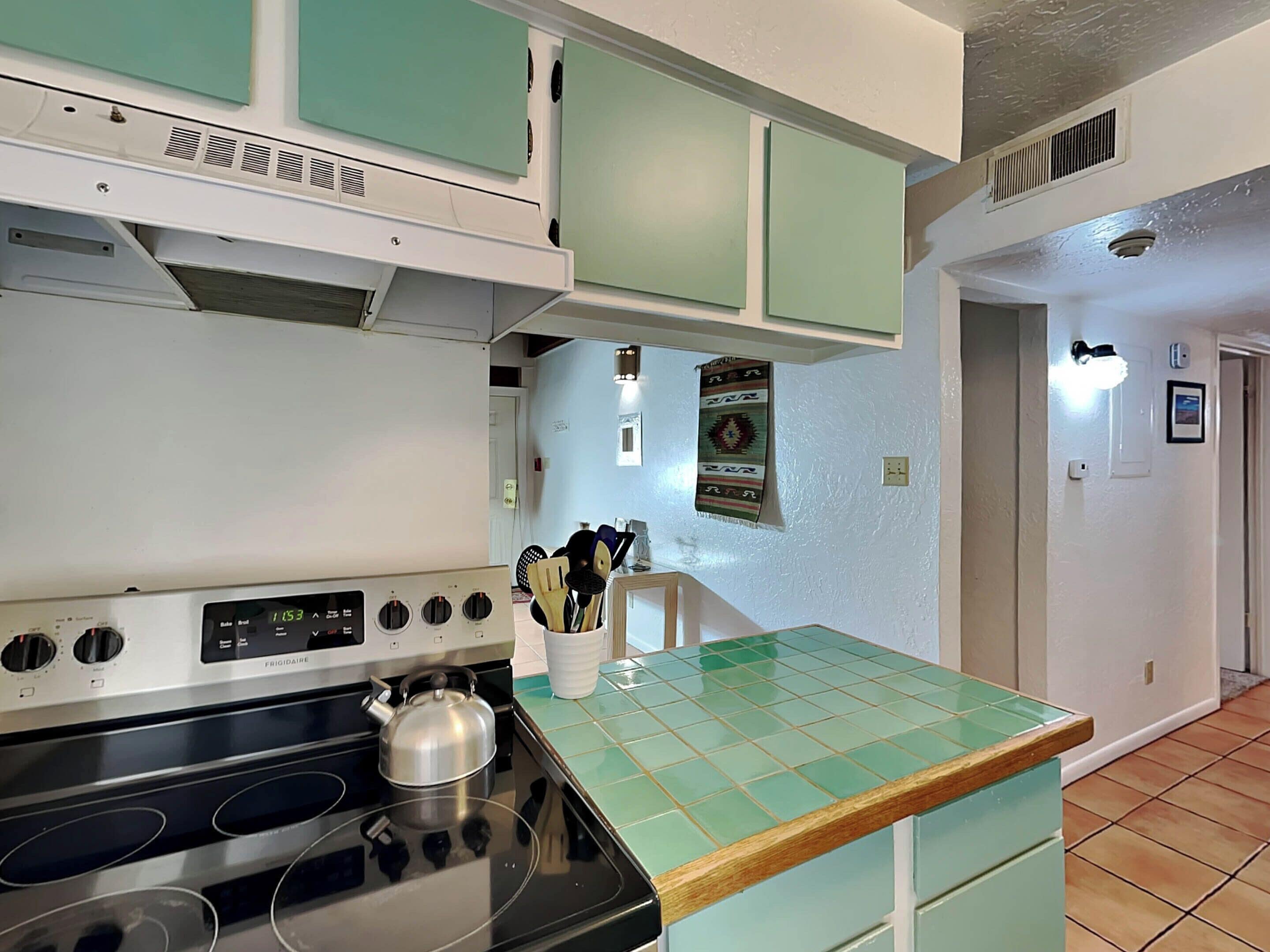 A kitchen with a stove, a kettle, and a cabinet with a green countertop.