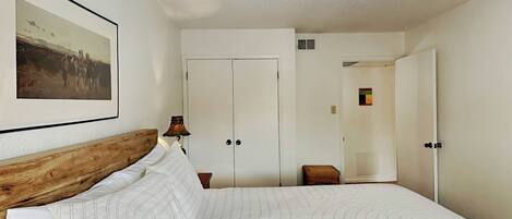 A bedroom with a large bed, a wooden headboard, a bedside lamp, a framed picture on the wall, and a closet with a door.