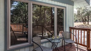 Une terrasse couverte avec deux chaises et une petite table, de grandes fenêtres et une vue sur une terrasse en bois et des arbres.