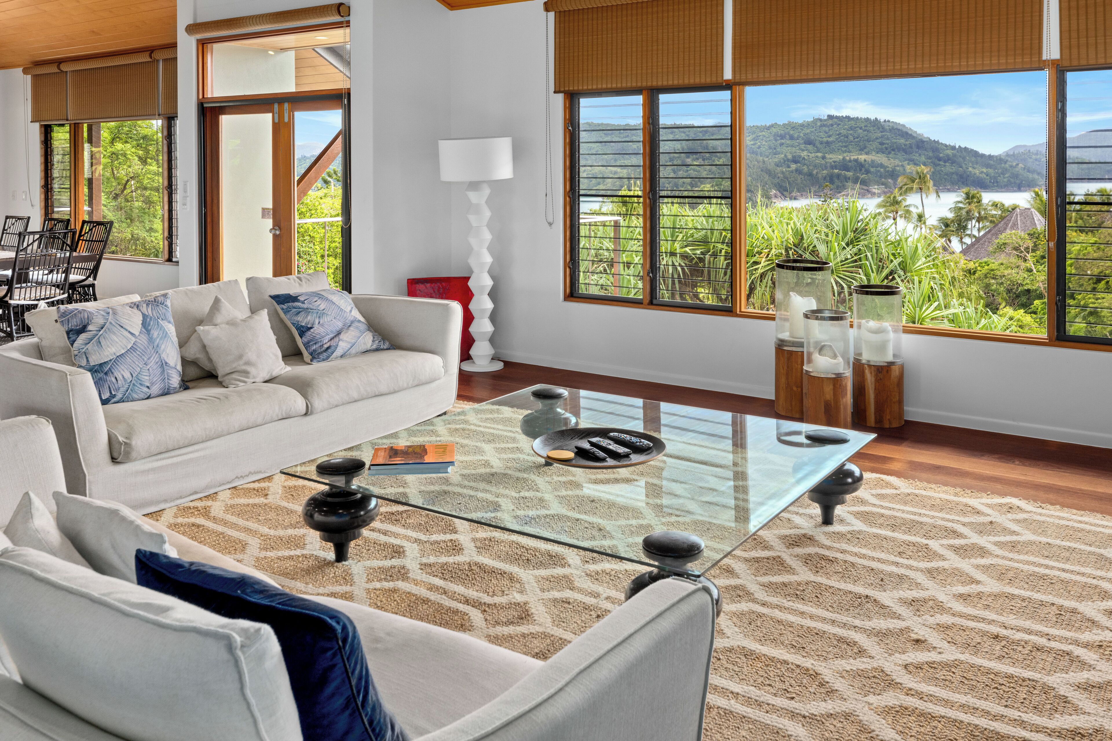 A modern living room with a glass coffee table, white sofa, and a view of a lake and mountains.