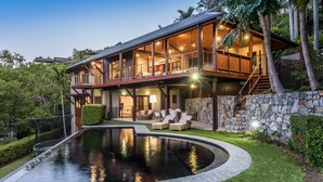 A two-story house with a large balcony, a swimming pool, and a stone wall. - Iluka Luxury House With Ocean Views On Half Acre With Pool And Two Golf Buggies (Whitsundays)