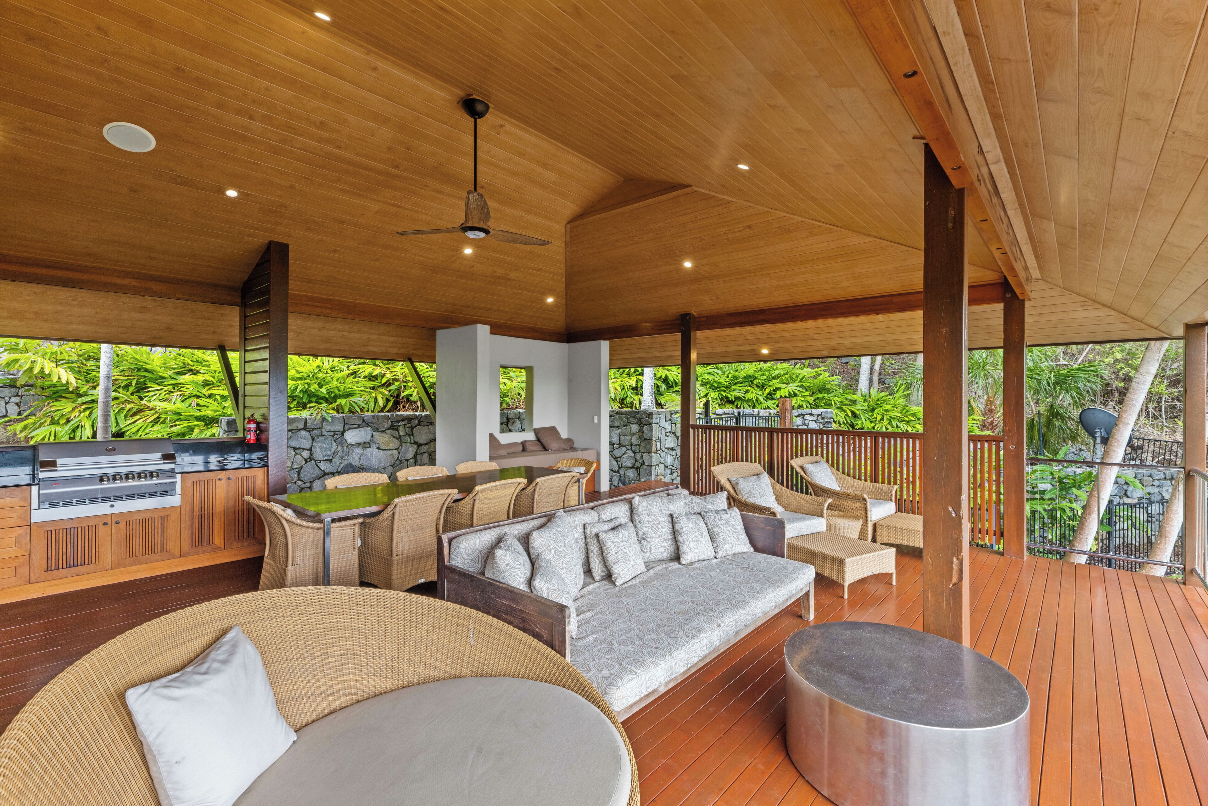 Un espacio al aire libre techado con terraza de madera, una parrilla incorporada, mobiliario de mimbre y un muro de piedra.
