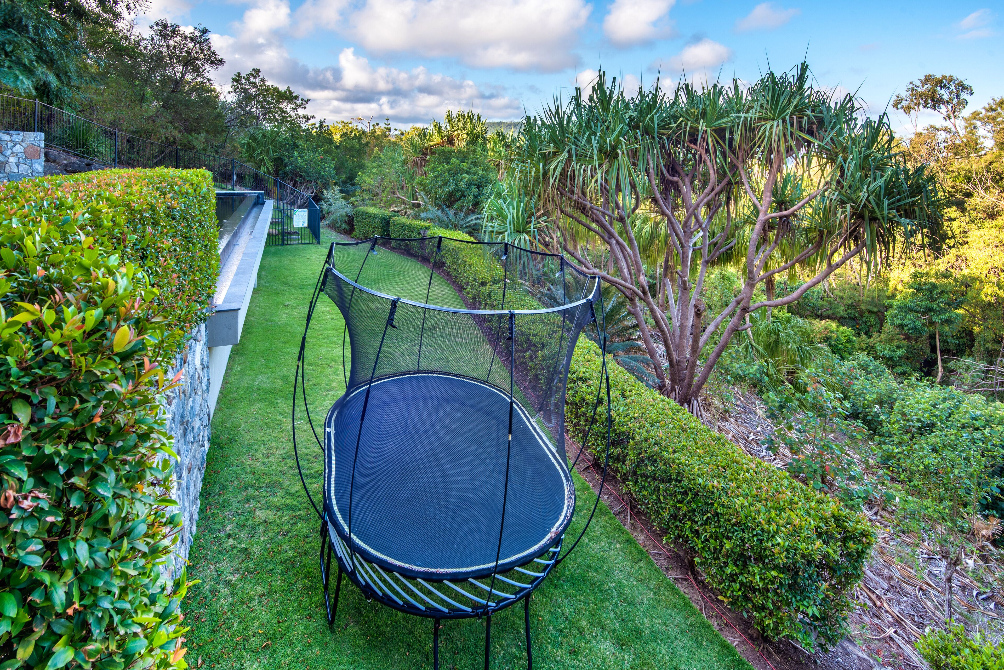 Un trampolín en un jardín exuberante con un muro de piedra y una valla de metal.