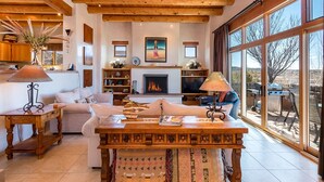 Un salon spacieux doté d’une cheminée, de poutres apparentes en bois et de grandes fenêtres offrant une vue sur l’extérieur.