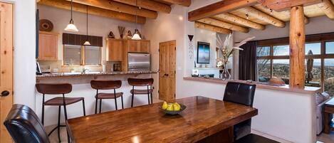 A spacious dining area with a wooden table and chairs, a kitchen in the background, and a view of the outdoors through large windows.