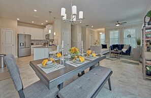 A modern kitchen with white cabinets, a stainless steel refrigerator, and a dining area set with a wooden table and benches. - Champions Gate Vista 1355 (Davenport)