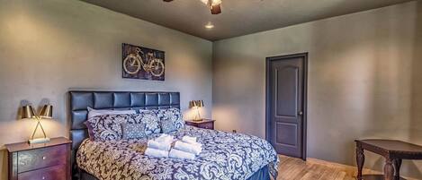 Une chambre avec un grand lit, un ventilateur de plafond, une œuvre d’art représentant un vélo accrochée au mur et un plancher en bois.