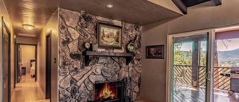 A stone fireplace with a fire, a wooden deck, and a view of the outdoors.