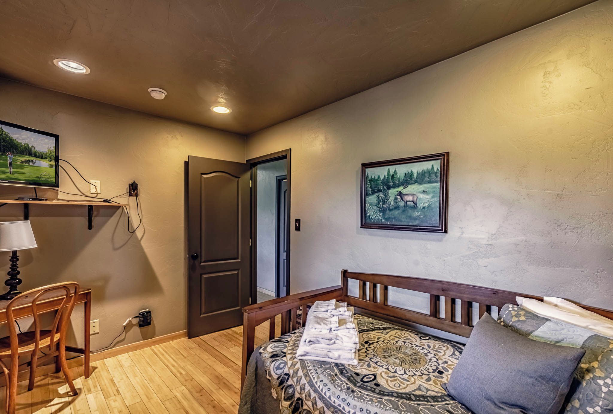 A room with a bed, a desk with a lamp, a chair, and a painting on the wall.
