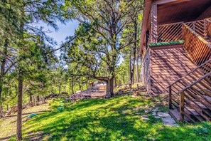 A wooden deck with a staircase leading to a covered area, surrounded by trees and a grassy area with outdoor furniture.
