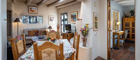 A cozy living room with a wooden dining table, chairs, and a sofa. There is a TV, a vase with flowers, and artwork on the walls.