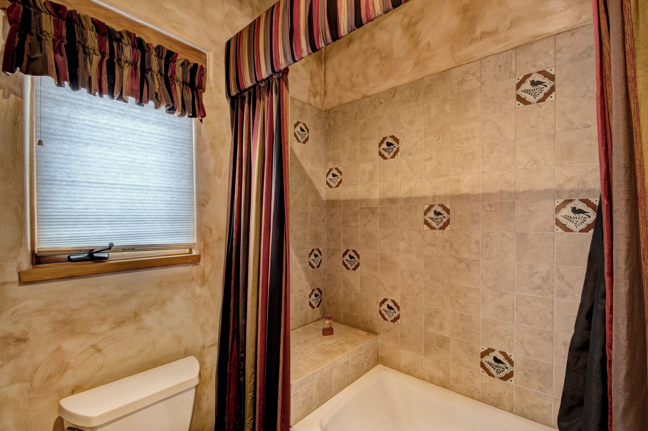 A bathroom with a toilet, a window with blinds, a shower with a tiled wall, and a bathtub.