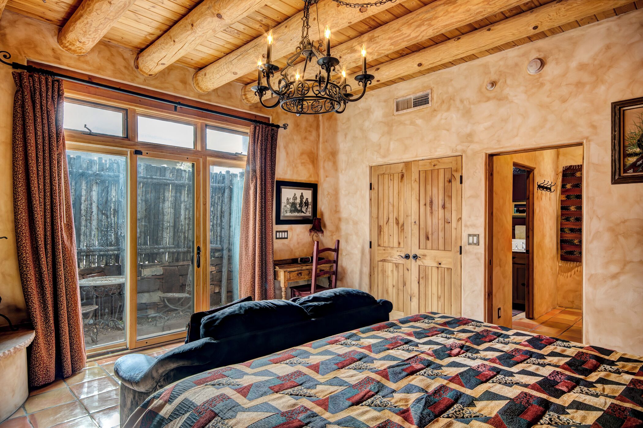 A bedroom with a large bed, wooden ceiling beams, a chandelier, a sliding glass door to a balcony, and a wooden wardrobe.