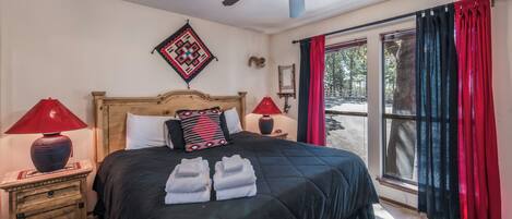 Un dormitorio con una cama, dos mesitas de noche, ventilador de techo, ventana con cortinas y vistas al exterior.