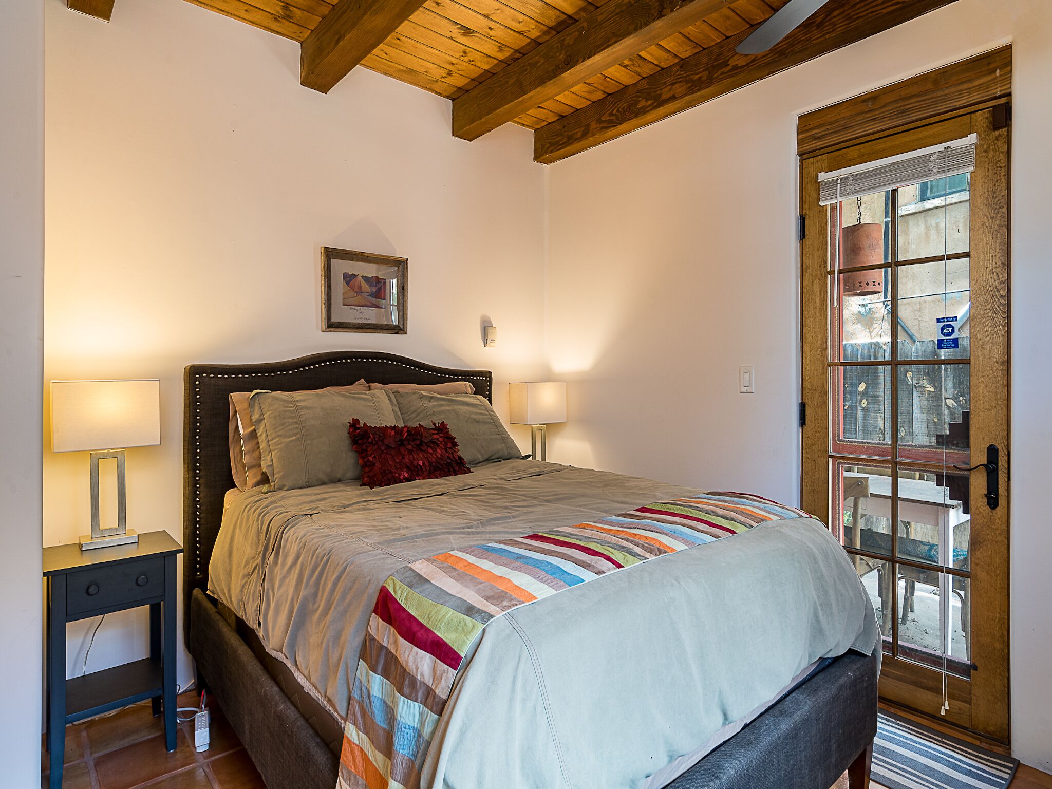A bedroom with a bed, two bedside lamps, a wooden ceiling, a door with a view, and a framed picture on the wall.