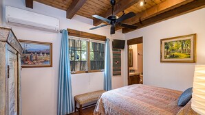 Ein Schlafzimmer mit Bett, Deckenventilator, einem Fenster mit Vorhängen, einem Bild an der Wand und einem sichtbaren Badezimmer durch eine offene Tür.