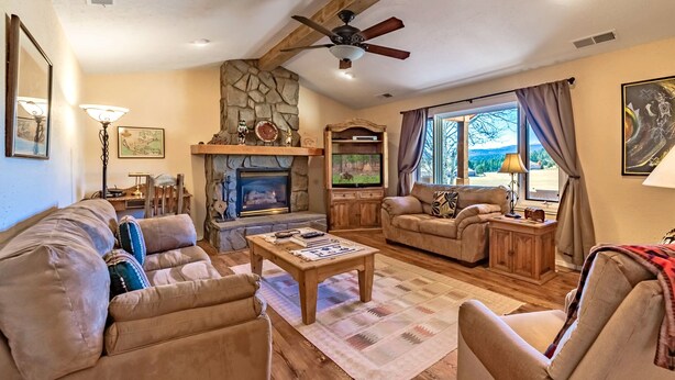 A cozy living room with a stone fireplace, wooden furniture, and a large window with curtains. - DB Mountain by Ruidoso Vacation Rentals (Ruidoso)