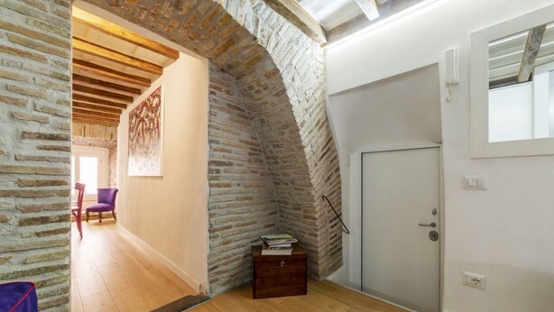 Quarto com piso de madeira, arco de tijolos, porta branca e um pequeno baú de madeira.