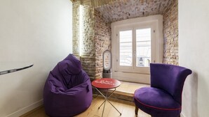 Quarto com parede de tijolos, uma porta, uma mesa pequena e duas cadeiras roxas.