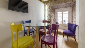 Uma área de refeições com mesa de vidro, cadeiras coloridas, televisão fixada na parede e uma parede de pedra com janela.
