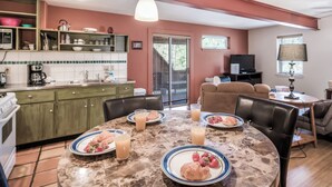 Un comedor con una mesa redonda dispuesta para el desayuno, una cocina al fondo y una sala de estar con un sofá y televisor.