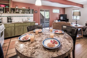 Ein Essbereich mit einem runden Tisch, gedeckt für das Frühstück, eine Küche im Hintergrund und ein Wohnzimmer mit Sofa und Fernseher.