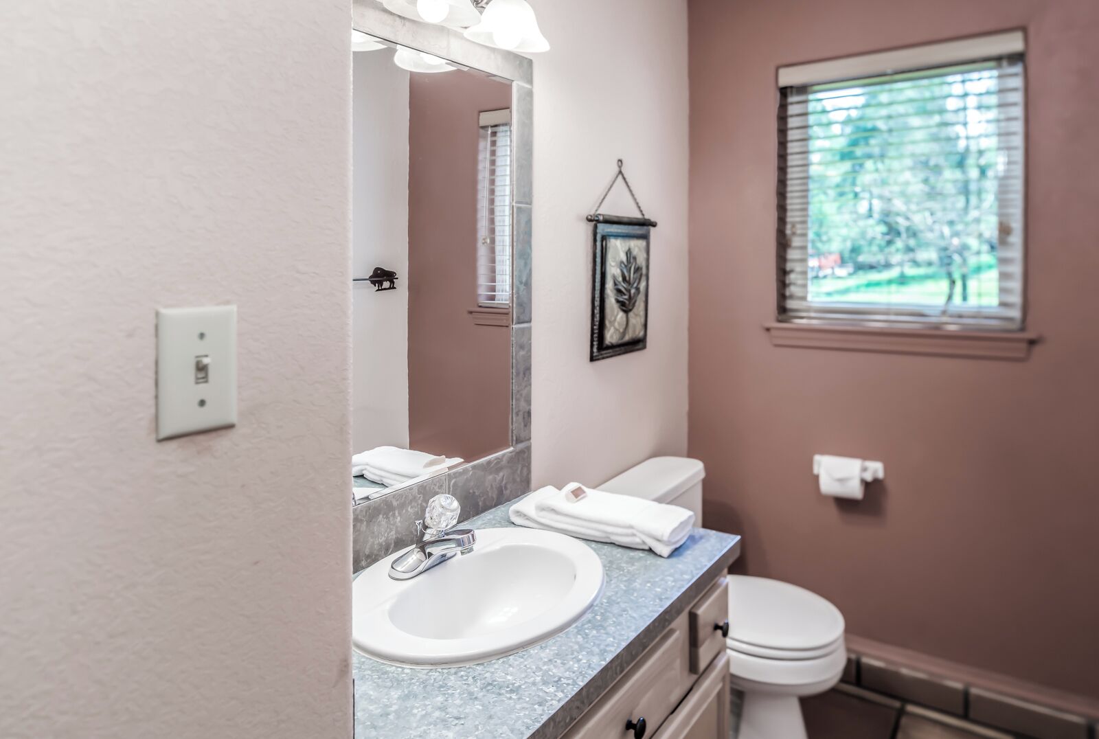 A bathroom with a white sink, a mirror, a toilet, and a window with blinds.