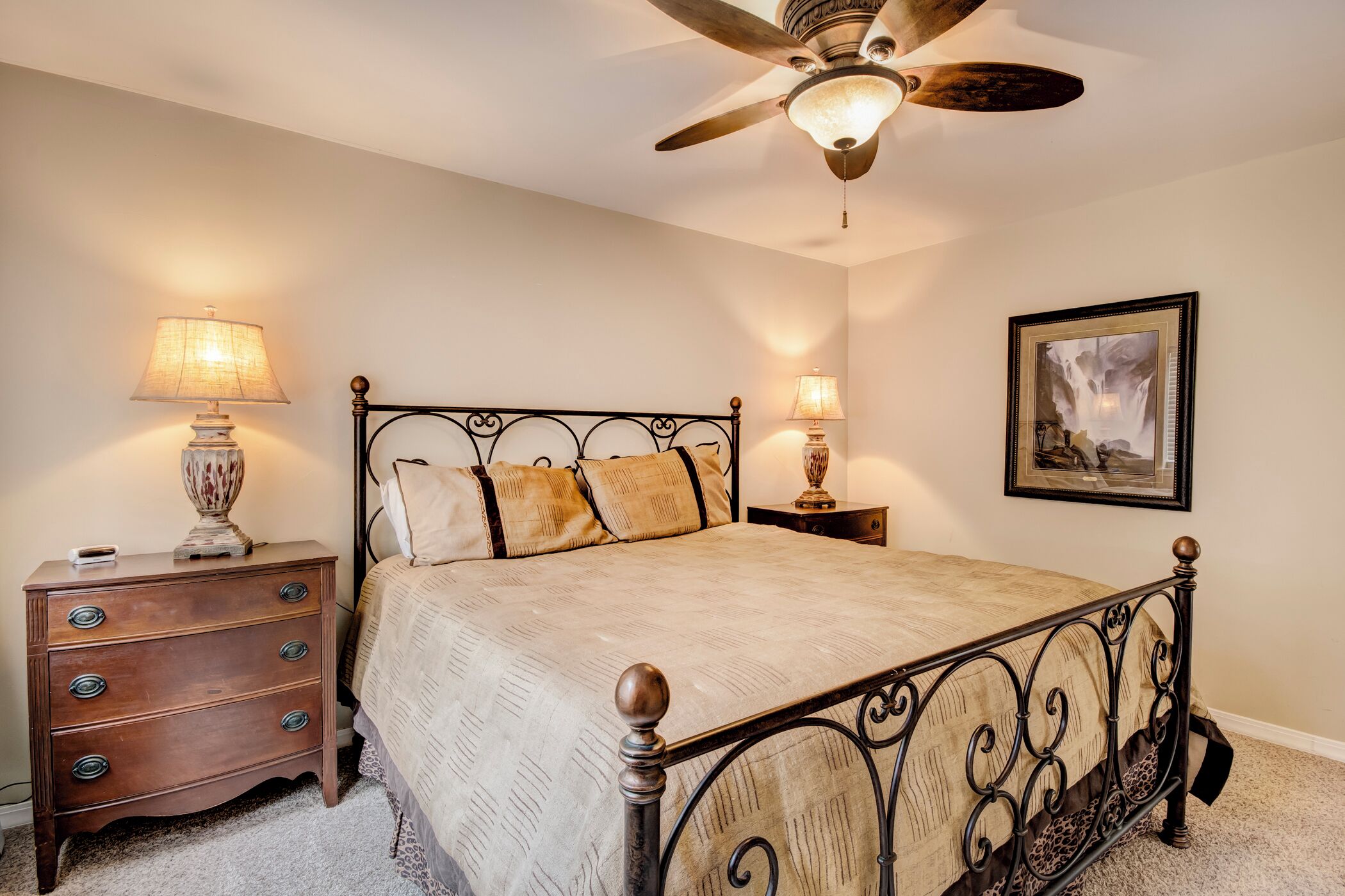 A bedroom with a bed, nightstands, lamps, a ceiling fan, and a framed picture on the wall.