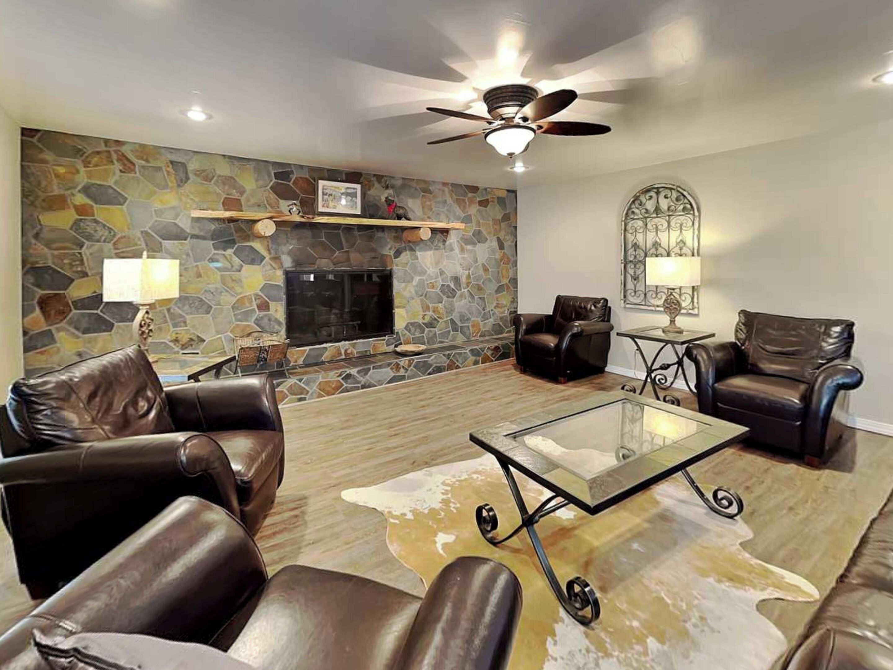 A living room with leather sofas, a glass coffee table, a stone fireplace, and a ceiling fan.