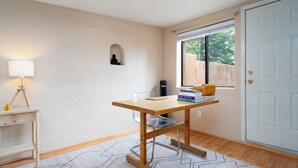 Een kamer met een houten tafel, een lamp, een klein plankje met boeken en een raam met uitzicht op groen.