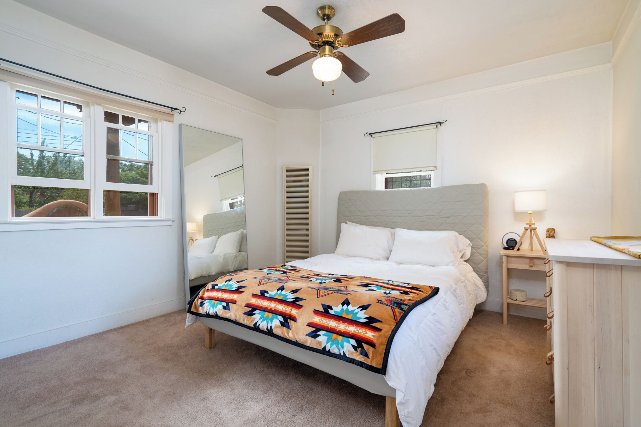 A bedroom with a bed, a ceiling fan, a bedside table with a lamp, a window with blinds, and a mirror.