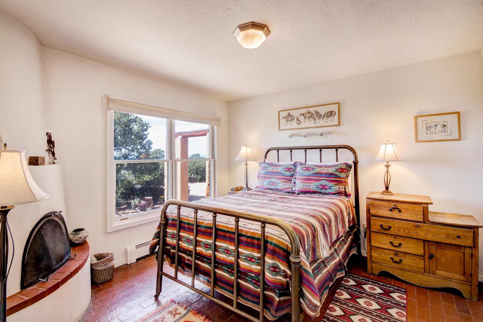 A bedroom with a bed, bedside lamps, a wooden dresser, a fireplace, and a window with a view of trees.
