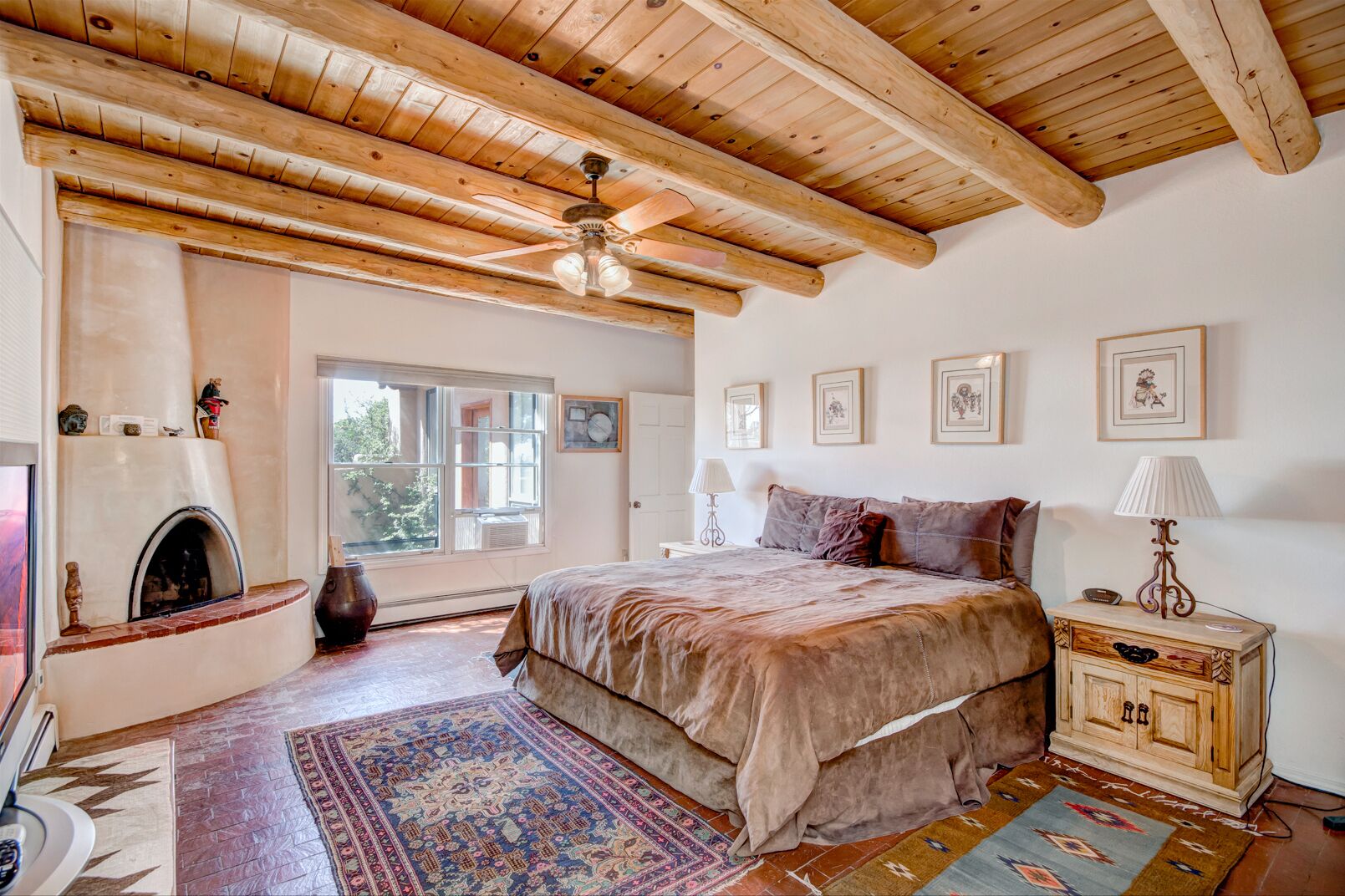 A bedroom with a large bed, wooden ceiling beams, a fireplace, and a window with a view of greenery.