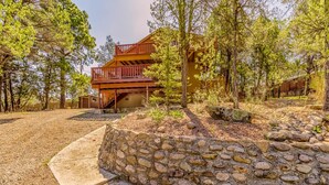 A two-story wooden cabin with a stone retaining wall and a gravel driveway. - Mockingbird Cabin by Ruidoso Vacation Rentals (Ruidoso Downs)