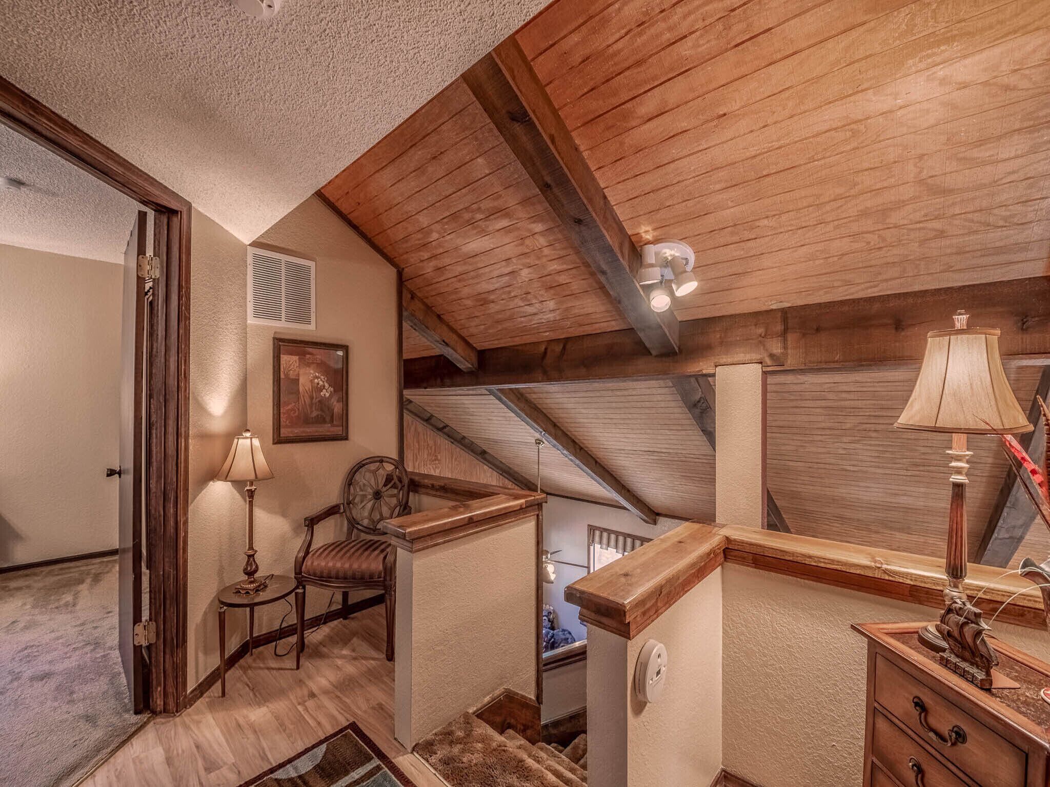 A loft-style bedroom with a sloped ceiling, wooden beams, a small seating area with a chair and table, and a lamp.