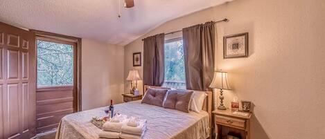 A hotel room with a large bed, a ceiling fan, a window with curtains, a bedside table with a lamp, and a framed picture on the wall.