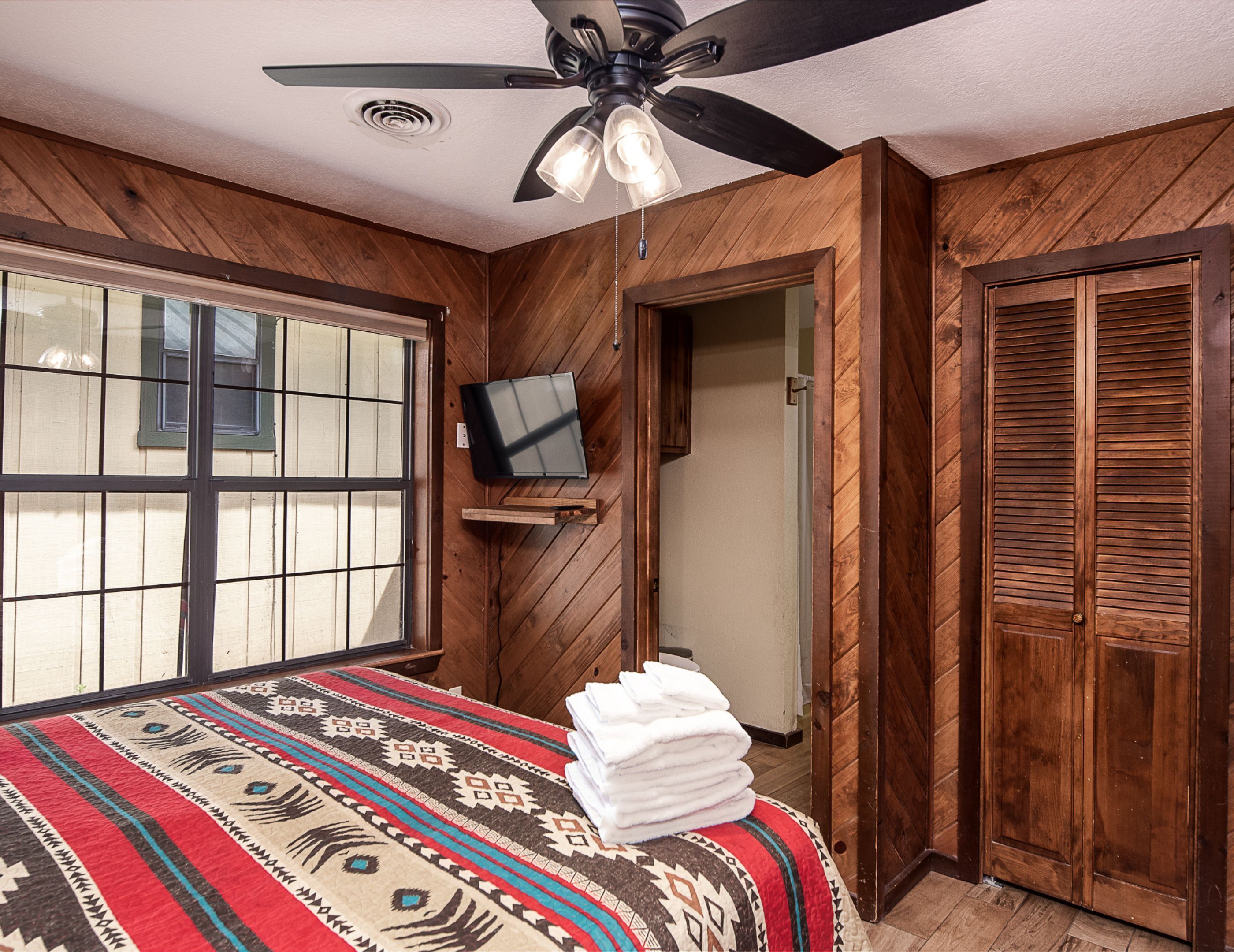 A bedroom with a bed, a ceiling fan, a TV mounted on the wall, and wooden paneling.